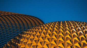 An architectural shot featuring a close-up view of two striking structures against a clear blue sky. The structure on the right is a golden, geometric dome made up of triangular patterns, while the structure on the left consists of a darker, grid-like design.