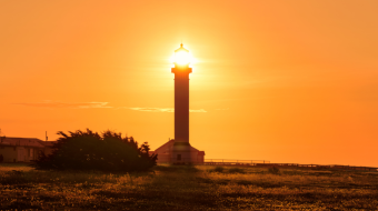 A tall lighthouse stands silhouetted against a vibrant orange and yellow sunset. The glowing sun sits directly above the lighthouse, making it appear as though the light is emanating from the tower. A field with sparse vegetation leads to the lighthouse.