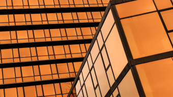 A modern architectural building with reflective glass windows displaying a warm, orange hue. The two structures intersect at an angle, showcasing sleek lines and a contemporary design, emphasizing the play of light and shadow on the glass surfaces.