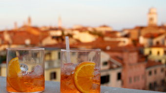 Two glasses of Aperol Spritz with orange slices and ice sit on a ledge, against a backdrop of a cityscape with blurred red-tiled rooftops and distant towers in warm sunset light. A straw is in one of the drinks.